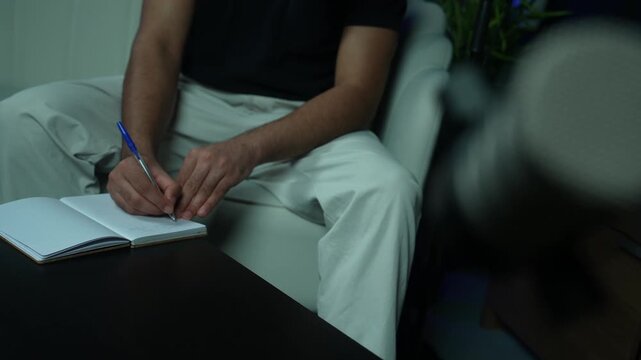 Close up of a man writing down notes in a notebook with a pen during a podcast recording session. Ideal for journalism, brainstorming, and creative media production concepts
