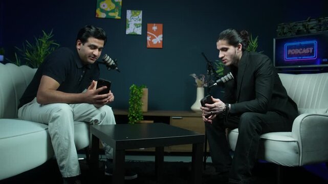 Wide shot of a male host and his guest both holding smartphones and discussing content during an interview in a modern podcast studio setup