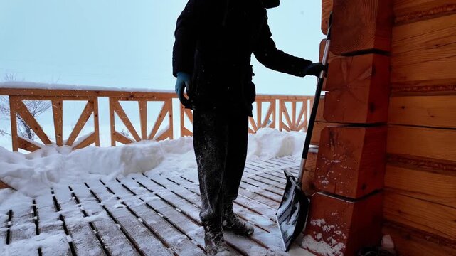 A man in dark winter clothing sets aside a wide snow shovel after clearing a snowy wooden terrace beside a log house during gentle snowfall.