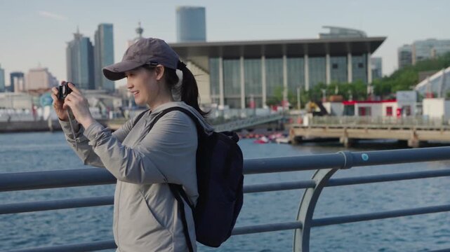 A female backpacker wearing casual outdoor clothing and a cap is taking photos with a compact camera at a waterfront urban harbor area.