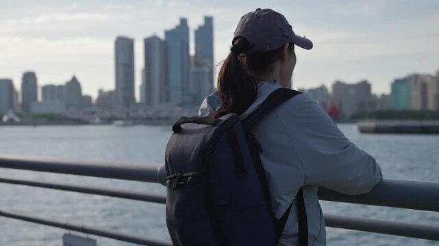 A female solo traveler in casual wear leans on a waterfront railing, overlooking a modern urban city skyline across the blue water.