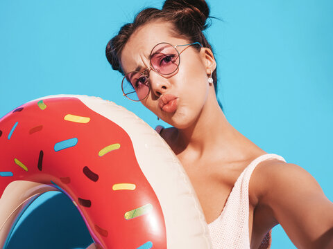 Young beautiful sexy smiling hipster woman in sunglasses. in summer clothes with donut lilo inflatable mattress.going crazy.Funny model isolated on blue.Taking selfie self portrait photos on phone