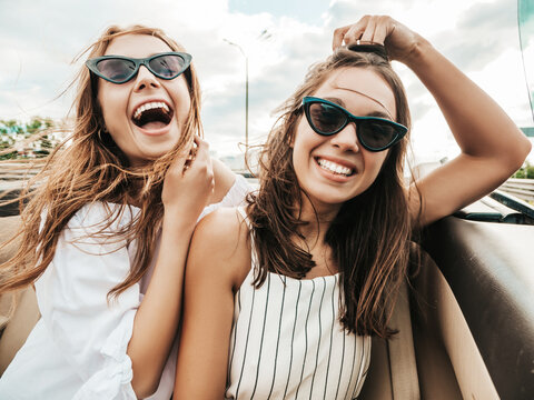 Portrait of two young beautiful and smiling hipster female in convertible car. Sexy carefree women driving cabriolet. Positive models riding and having fun in sunglasses outdoors.Enjoying summer days
