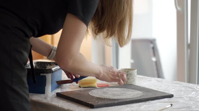 Woman applies white plaster texture to canvas panel with spatula on covered table. DIY textured art, handmade decor and creative process concept.