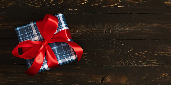 Horizontal Banner of Plaid Wrapped Gift Box Tied with Vibrant Red Ribbon Bow on Dark Wooden Background with Copyspace, Creative Top View Flat Lay for Christmas, Father's Day or Birthday