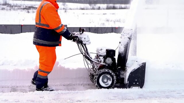 Snowblower operator in bright orange suit clears snow. Snow plows forcefully ejects thick snow into air. Operator controls machine with steady, focused posture