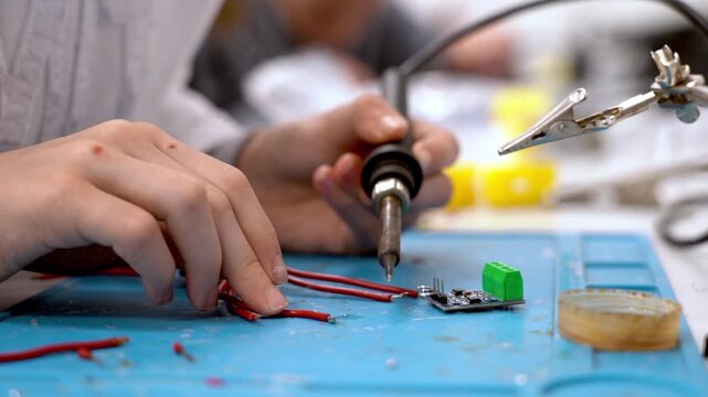 Hands work with soldering iron and wires. Focus on precision and technical skill. Blue mat holds tools and components. Hands steady, showing concentration. Perfect for electronics or DIY projects