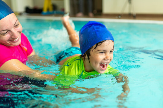 Swimming lesson. Joyful boy lies on his stomach kicking legs through water while a smiling female instructor supports his body, practicing horizontal float position  