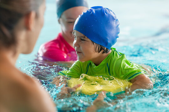 Boy holds a kickboard in an indoor pool, attentively listening to a female instructor during a structured aquatic lesson, with a second instructor visible in the background.