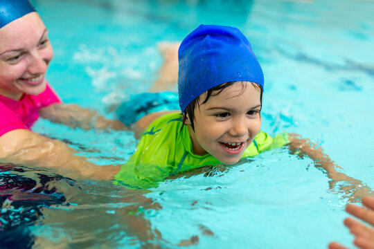 Laughing boy lies flat on water surface supported by a female instructor, practicing front glide technique in an indoor pool during a structured early childhood swimming lesson.
