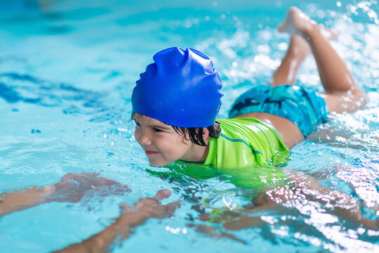Smiling boy in a blue swim cap and green vest swims alongside a female instructor in a turquoise pool, gaining water confidence through guided aquatic training.