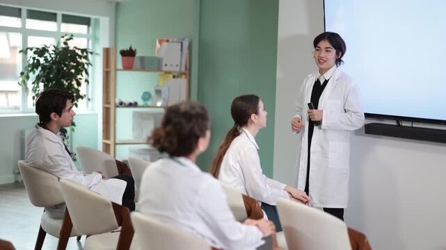 Young female doctor in medical gown with remote control gives presentation to colleagues doctors in conference room
