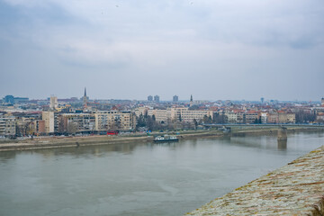 Obraz premium A panoramic view of Novi Sad, Serbia, showcasing the city skyline along the Danube River. Buildings line the riverbank under a cloudy sky.