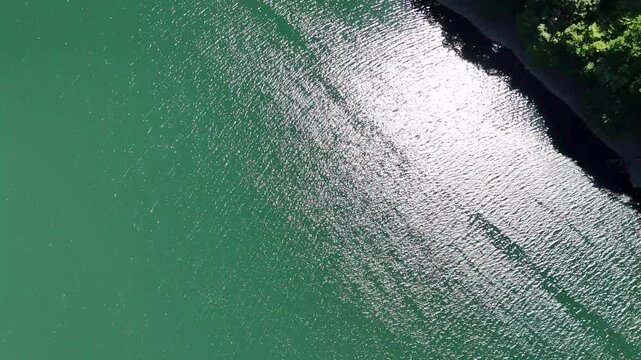 Shimmering water reflects bright sunlight. Ripples dance across calm, greenish surface. Sunlight glints vividly against gentle waves. Riverbank edges frame tranquil, natural scene