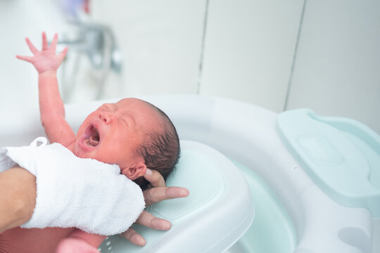 Close-up of a crying newborn baby being bathed by a parent in a plastic support tub with a white towel.