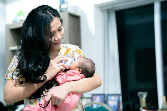 Smiling Asian mother holding and bottle-feeding her newborn baby with love and care at home.