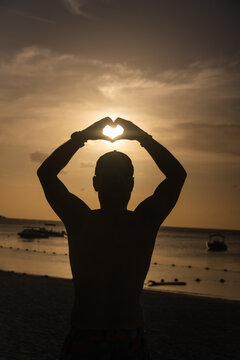 silhouette of a man on the beach forming a heart with his hands catching the sun at sunset