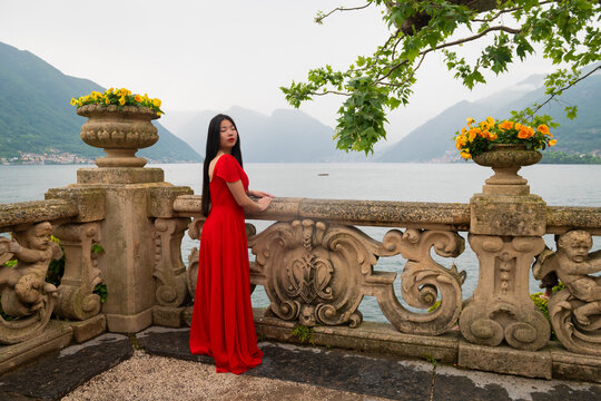 art portrait of asian young woman in red dress near stone balustrade on terrace at lake como, italy