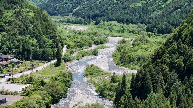 River winds through lush green mountains. Rocks and gravel bed the rushing stream. Trees line both banks, dense and tall. Village sits quietly near the water's edge. Scenic view invites calm