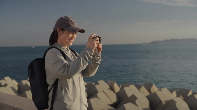 A smiling young female hiker with a backpack and cap uses a compact camera to take seascape photos at a coastal breakwater during daytime.