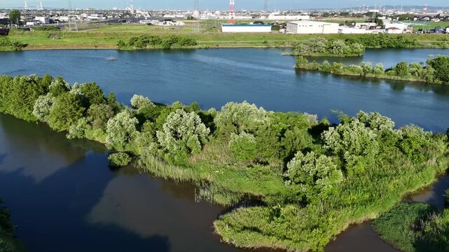 Aerial view of a winding river through greenery. Trees frame the water's edge, softly swaying. Islands rise from the calm, reflecting the sky. Distant buildings hint at human presence