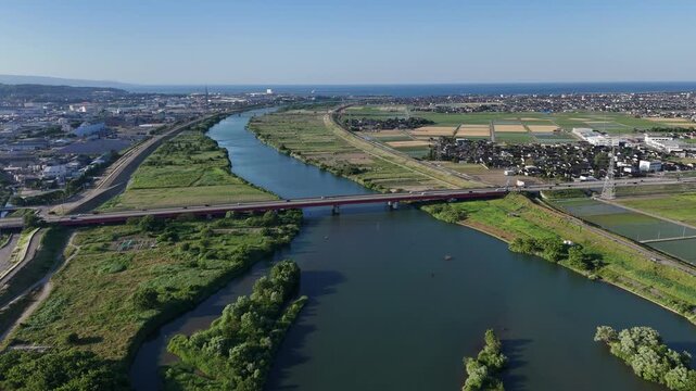Aerial view of river flowing through green landscape. Bridge spans water, connecting two sides. Vibrant green fields surround the riverbank. Clear sky above with distant city skyline