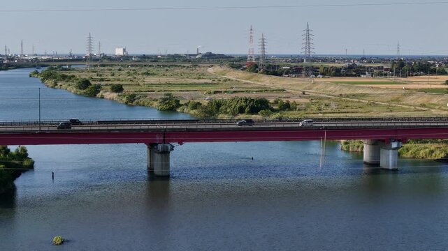 Red bridge spans calm river under clear sky. Concrete pillars anchor structure with steady grace. Vehicles glide smoothly across span in quiet motion. Power lines stretch distant