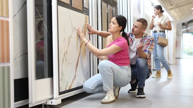 Enthusiastic young interracial couple browsing together technical and natural stone tile samples on display board in home improvement store, comparing colors and textures before purchasing. High