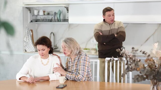 Old woman asking livid middle-aged woman for forgiveness during domestic quarrel in the kitchen