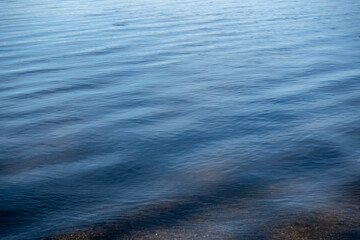 Obraz premium Gentle ripples on the surface of clear blue water, showing a pebble-strewn bottom