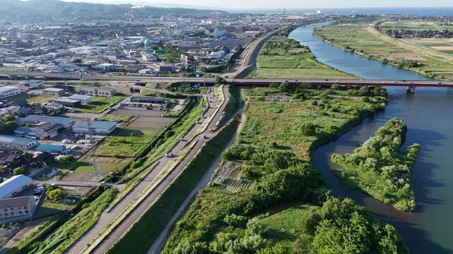 Aerial view of river beside town and highway. Bridge spans water, linking traffic and land. Green banks line calm riverbed, dotted with trees. Road curves past fields, leading to urban center