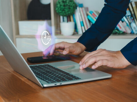 Hands typing on laptop keyboard with smartphone nearby and holographic clock icon symbolizing productivity and time management in home office setting