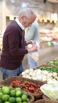 Senior man is standing in a supermarket and choosing a white turnip to make a salad. Elderly buyer puts the vegetables in a bag to weigh before buying. High quality 4k footage