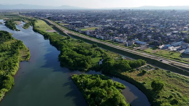 Aerial view of winding river with lush green banks. City sprawls beside the river, separated by a highway. Sunlight glints softly off the water's surface. No people visible, nature