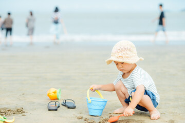 A cute 3-year-old Chinese boy wearing a straw hat while crouching and digging sand with a toy shovel and bucket at Hetou Beach, Banbianshan, Xiangshan, Ningbo, Zhejiang, China © sweetriver