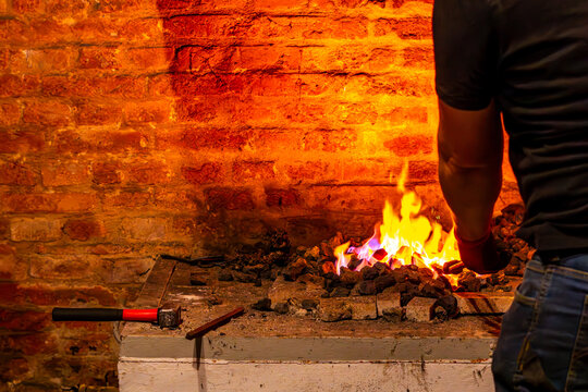 A man viewed from behind works at a glowing blacksmith forge with burning coal, its intense orange flames illuminating an old red brick wall and nearby tools like a hammer and chisel.
