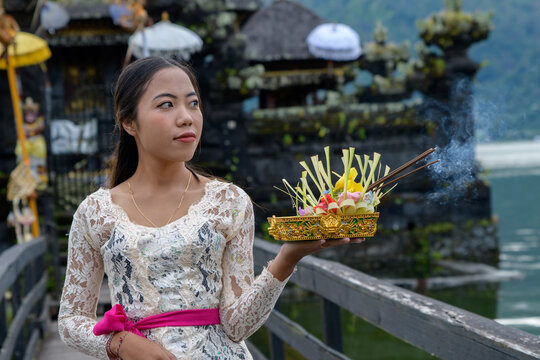 Balinese woman in white lace kebaya with offering tray looking up at Pura by mountain lake