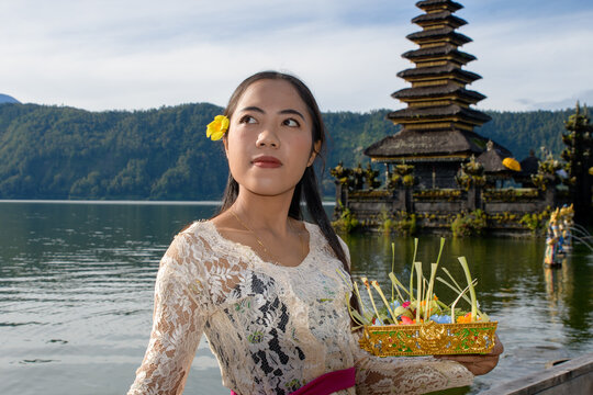 Young Balinese woman in white kebaya carrying ritual offering at temple with lake and mountain backdrop