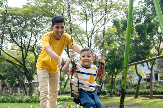 Happy Asian father pushing his cute little son on a swing at the playground