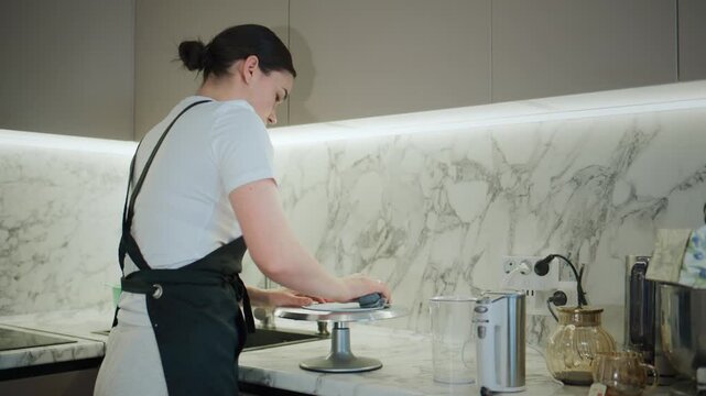 Young woman in apron cleans countertop with focused attention. Modern kitchen with marble backsplash and warm lighting. She appears calm and dedicated to her task. Footage ideal for food service