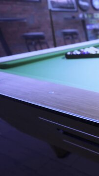 The scene shows a pool table in a bar. Balls are arranged on the table waiting to be played. The setting has soft lighting, and the atmosphere feels social and lively.