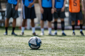 Fototapeta premium Close up of soccer ball on green artificial turf pitch with blurred players background, football match training session concept before kickoff at sports stadium
