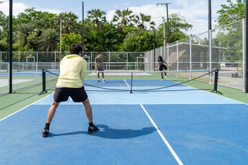 Naklejka premium Group of young multiethnic friends playing pickleball doubles match on outdoor court. Active athletes holding paddle rackets during padel tennis game training session