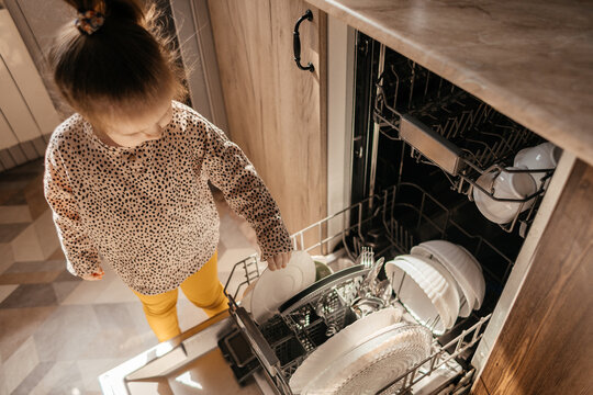 Little girl unloading clean plates from dishwasher in kitchen