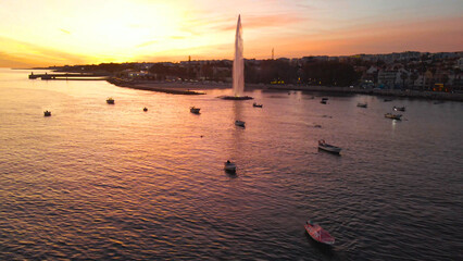 Oeiras fountain stands tall, spraying water into the golden sky at sunset, while numerous small boats anchor peacefully in the tranquil bay along the Portuguese coastline © Dark Artistic