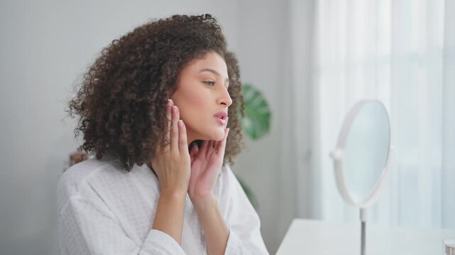 Latina woman looking in the mirror while applying lotion to her face. 
