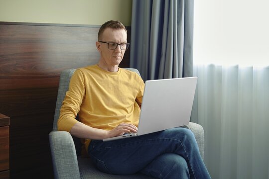 A man sits in a comfortable chair, focused on his laptop in a bright room. Soft natural light streams through the curtains as he engages in his tasks, showcasing a productive atmosphere