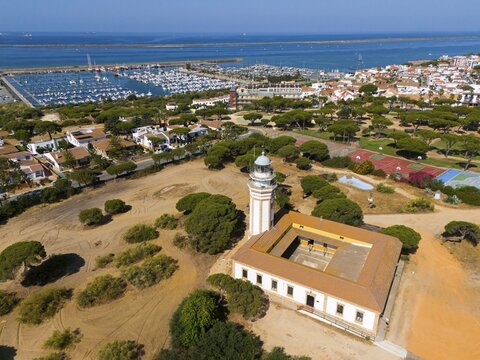A lighthouse overlooking a coastal town and a marina, surrounded by trees under a sunny sky, aerial view, lighthouse, Mazag&oacute;n, Mazagon, Cadiz province, Cadiz, Costa de la Luz, Andalucia, Spain