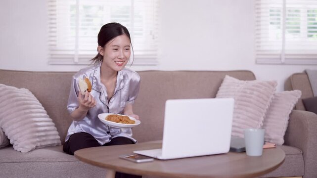 Asian woman enjoy breakfast snack eating cookie food working from home on laptop computer relaxing on sofa living room morning routine home office remote work