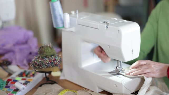 Professional female seamstress working at sewing machine, stitching natural linen fabric with focus and precision at table in workshop, cropped shot